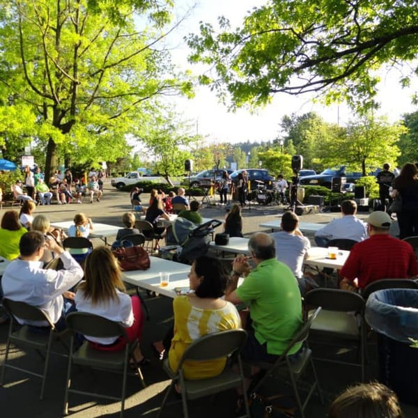 People seated at tables outdoors, watching a band perform under green trees at Rice Thai Cookery.