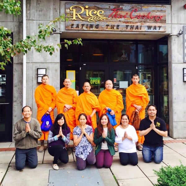 Monks in orange robes and people kneeling with hands pressed together outside Rice Thai Cookery.